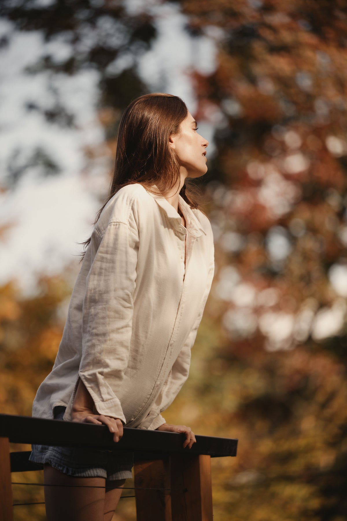 Woman in an oversized white triple gauze easy shirt leaning on a railing among autumn trees